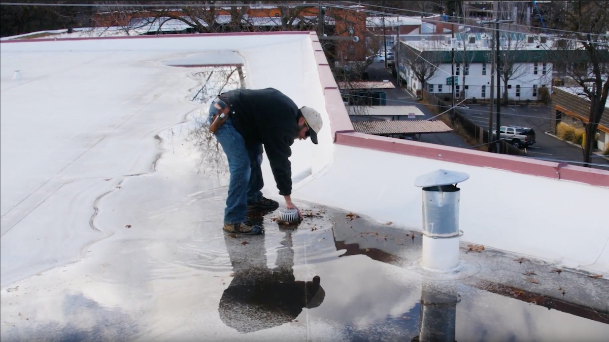 Roof Reporter Clearing Debris From Drain During Flat Roof Inspection To Relieve Ponding